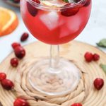 A glass of red drink with ice cubes, cranberries, a lemon slice, and mint leaves, placed on a wooden coaster with scattered cranberries around it—this refreshing mocktail with cranberry juice is the perfect fall mocktail to enjoy as the seasons change.