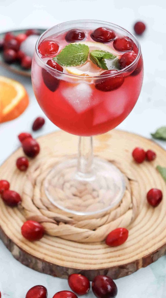 A glass of red drink with ice cubes, cranberries, a lemon slice, and mint leaves, placed on a wooden coaster with scattered cranberries around it—this refreshing mocktail with cranberry juice is the perfect fall mocktail to enjoy as the seasons change.