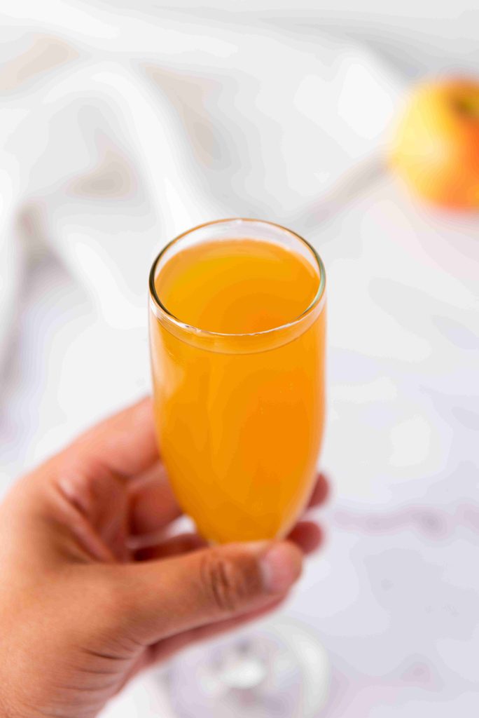 A hand holding a glass of orange-colored drink, possibly a fall mimosa, with a blurred background and an apple visible in the top right corner—perfect inspiration for your next fall mimosa recipe.