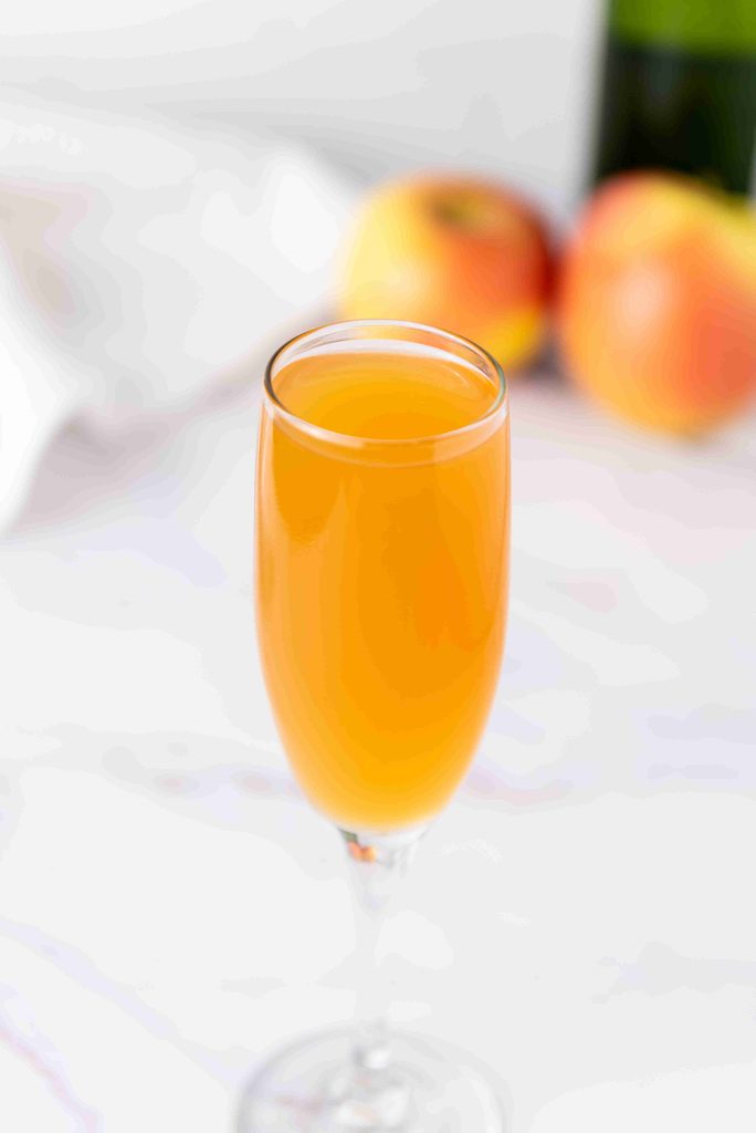 A glass of orange-colored beverage, likely a fall mimosa, sits on a white surface with blurred apples and a green bottle in the background.