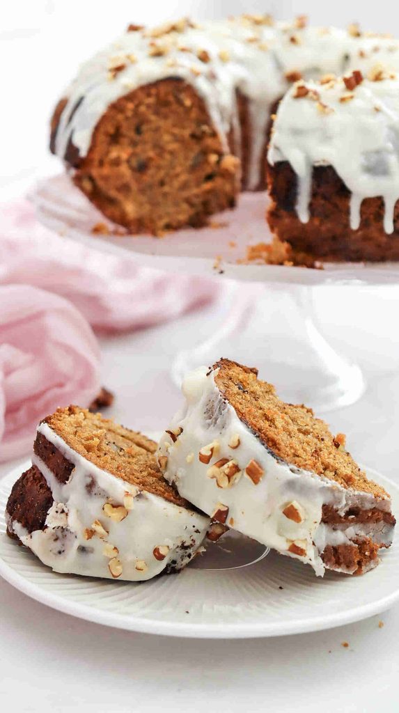 Two slices of fall bundt cake with white icing and chopped nuts rest on a plate, with the remaining cake on a stand in the background—an inviting treat perfect for any fall bundt cake recipe enthusiast.