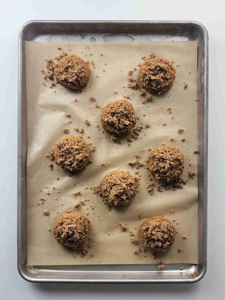 A baking tray lined with parchment paper holds eight unbaked pumpkin cookies with icing, shaped into dough balls with crumbly bits scattered around them.