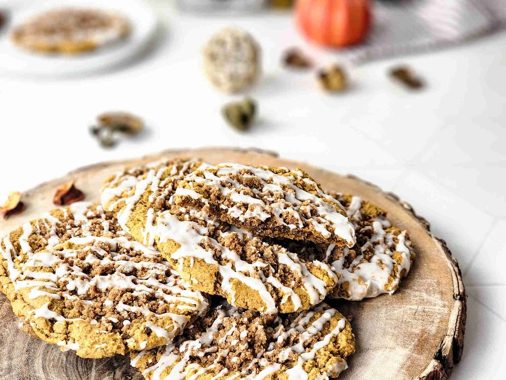Three large pumpkin coffee cake cookies with crumbly topping and white icing drizzle sit on a wooden platter. The background is bright and out of focus, with hints of autumn decor and blurred objects.