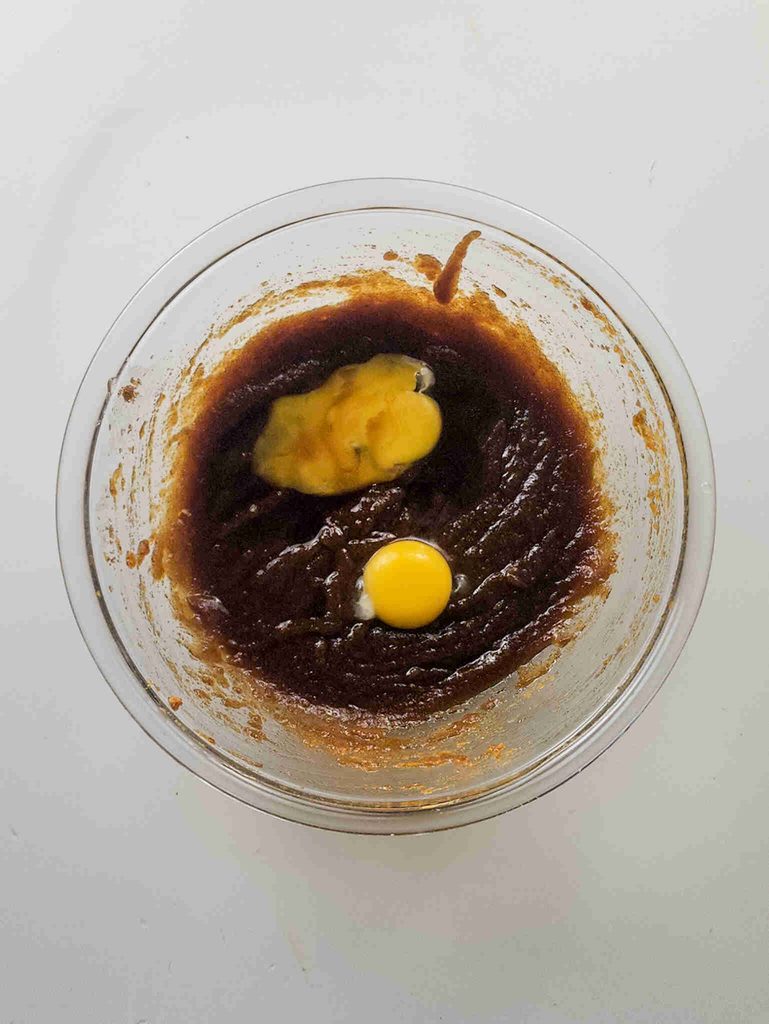A glass bowl containing dark brown batter for pumpkin cookies with icing, with a whole raw egg and a dollop of yellowish mixture on top, viewed from above on a white surface.