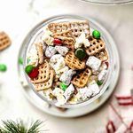 A glass bowl filled with reindeer munch, a snack mix of pretzels, popcorn, and red and green candies dusted with powdered sugar.