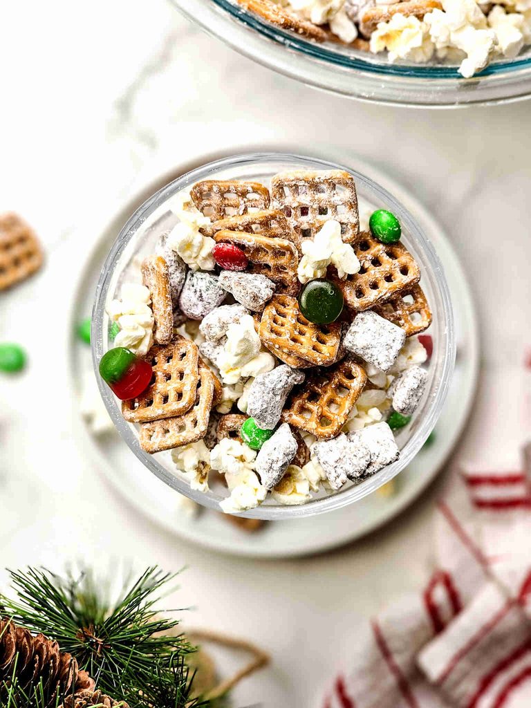 A glass bowl filled with reindeer munch, a snack mix of pretzels, popcorn, and red and green candies dusted with powdered sugar.