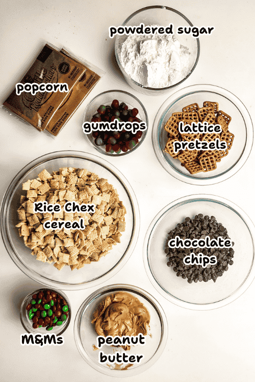 Overhead view of ingredients for a Christmas puppy chow reindeer munch mix: popcorn, powdered sugar, gumdrops, lattice pretzels, Rice Chex cereal, chocolate chips, M&Ms, and peanut butter, all labeled.