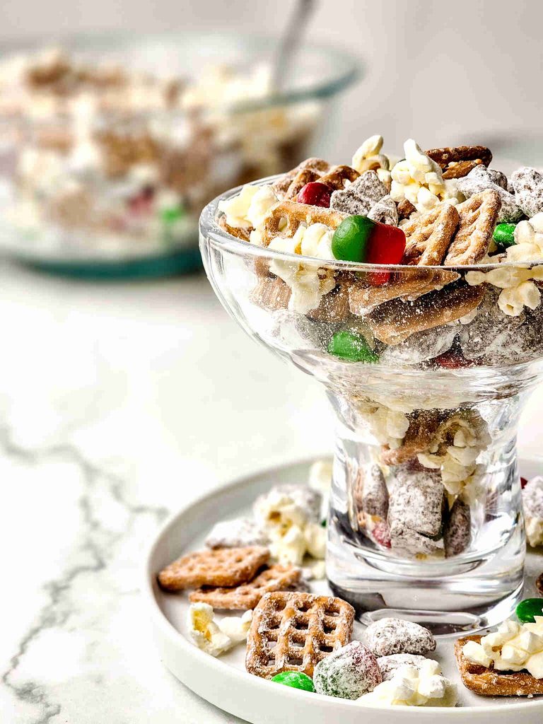 A glass dessert cup filled with reindeer munch, a snack mix of pretzels, popcorn, and colorful candies coated in powdered sugar.