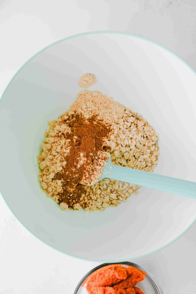 A mixing bowl containing rolled oats, protein powder, and cinnamon for high protein pumpkin overnight oats, with a silicone spatula stirring the ingredients and a small bowl of pumpkin purée nearby.