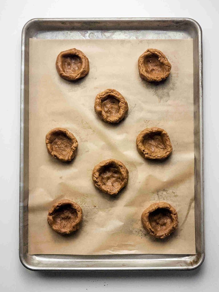 Eight unbaked Pumpkin Pie Cookies with indents in the center are arranged on a parchment-lined baking sheet, ready for filling with custard filling or baking.