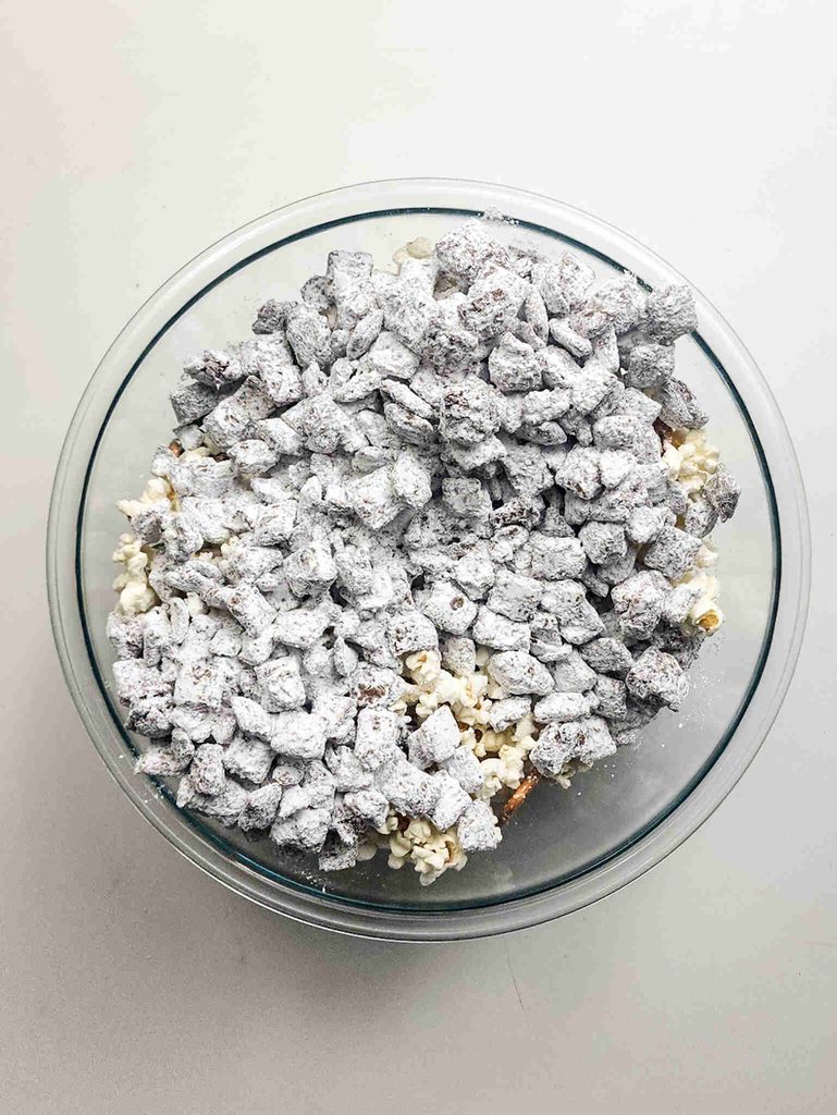 A clear glass bowl filled with a mixture of powdered sugar-coated chocolate cereal pieces and popcorn, commonly known as Christmas puppy chow.