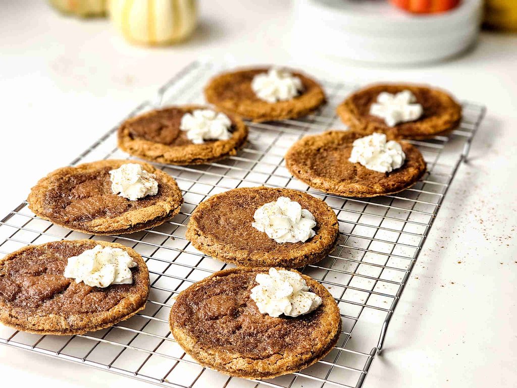 Eight round Pumpkin Pie Cookies topped with dollops of whipped cream are cooling on a wire rack, with small pumpkins and kitchen items blurred in the background.