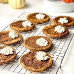 Eight round Pumpkin Pie Cookies topped with dollops of whipped cream are cooling on a wire rack, with small pumpkins and kitchen items blurred in the background.