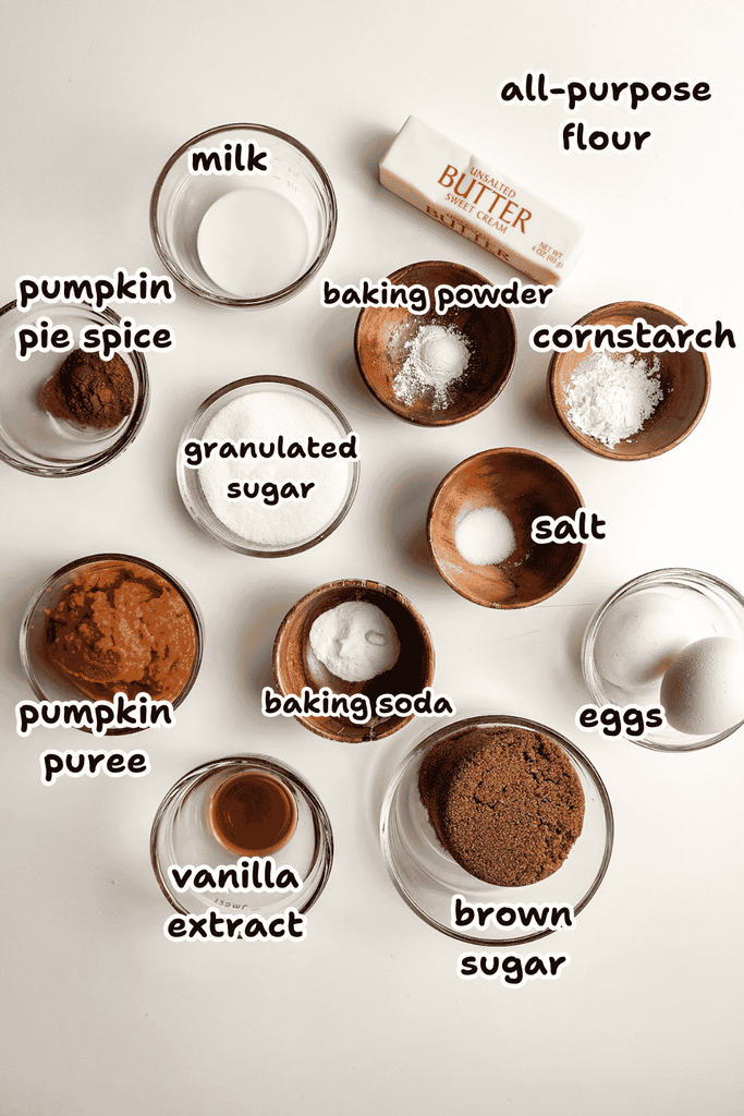 Twelve labeled ingredients for baking Pumpkin Pie Cookies are arranged on a white surface, including flour, butter, pumpkin puree, spices, and more—perfect for cookies with a whipped cream or custard filling.