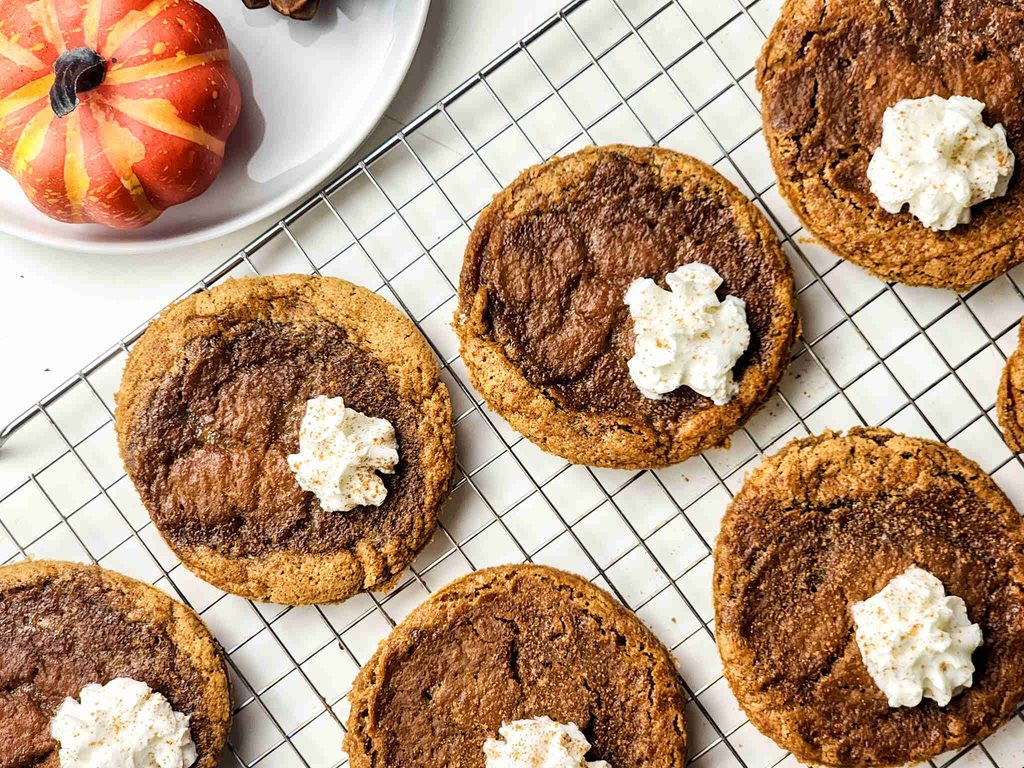 Pumpkin Pie Cookies with whipped cream dollops are cooling on a wire rack. A decorative pumpkin and a white plate are partially visible in the top left corner.
