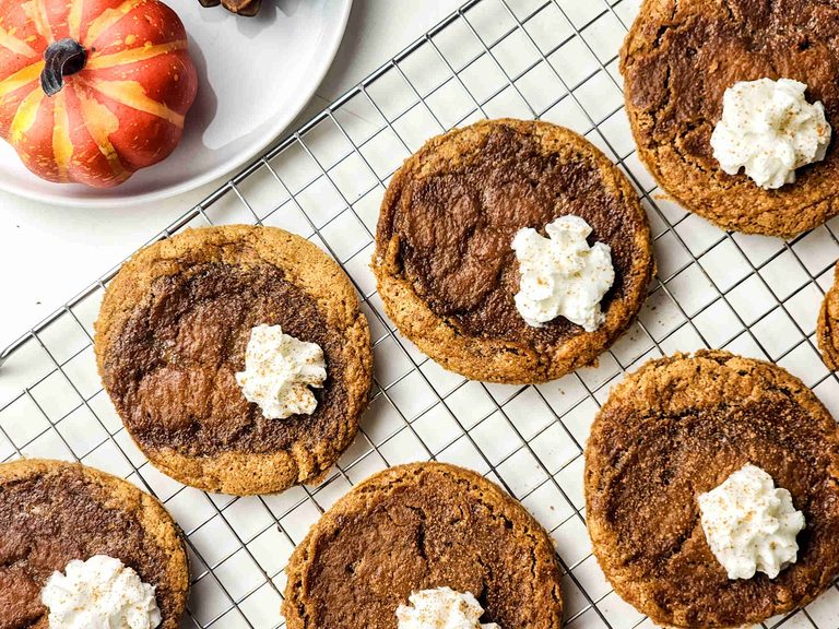 Pumpkin Pie Cookies with whipped cream dollops are cooling on a wire rack. A decorative pumpkin and a white plate are partially visible in the top left corner.
