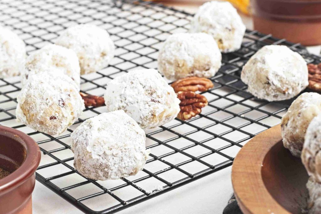 Powdered sugar-coated pumpkin snowball cookies cool on a black wire rack, with pecan halves scattered underneath and ceramic dishes nearby.
