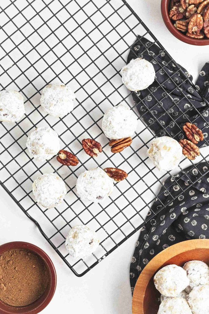 Powdered sugar-coated pumpkin snowball cookies rest on a cooling rack, with scattered pecans nearby. Bowls of pecans and cocoa powder sit on the white surface beside a patterned cloth and wooden bowl.