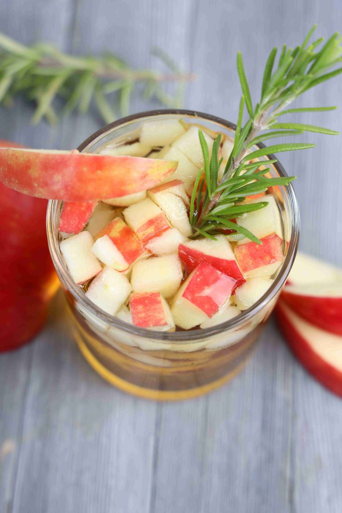 An apple cider Thanksgiving cocktail in a glass filled with diced apples and garnished with a sprig of rosemary.