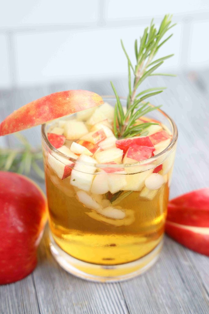 A glass of apple cider garnished with diced apples and a sprig of rosemary, perfect as a Thanksgiving cocktail.