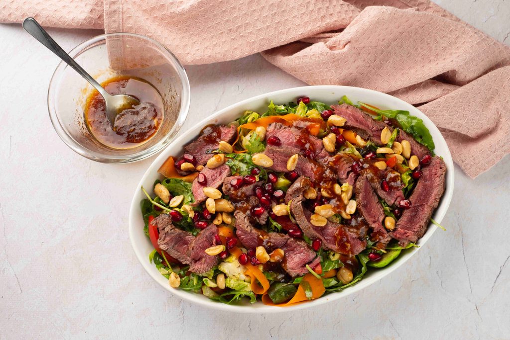 A fall steak salad of sliced beef, peanuts, pomegranate seeds, and greens in an oval bowl, with a side bowl of dressing.