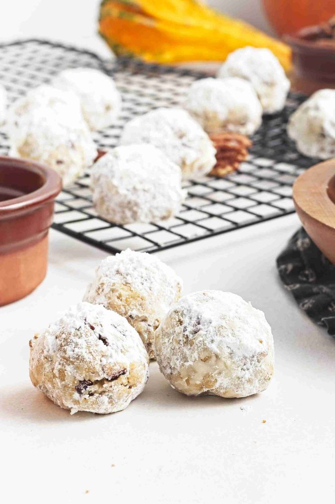 Three powdered sugar-covered pumpkin snowball cookies sit in front of a cooling rack with more cookies. A small brown bowl and scattered pecans are visible, with an orange item blurred in the background.