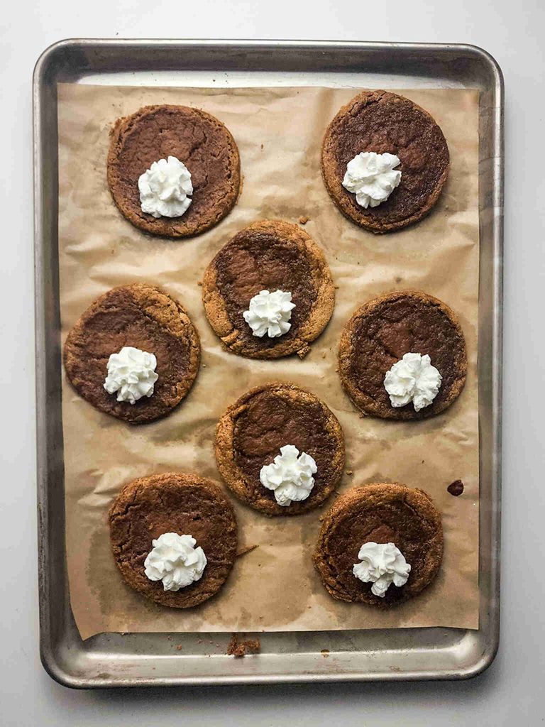 Eight pumpkin pie cookies topped with whipped cream are arranged on a parchment-lined baking sheet, placed on a white surface.