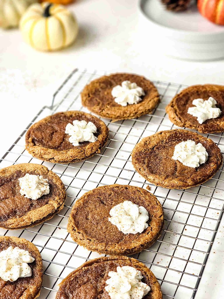 Eight Pumpkin Pie Cookies, half topped with whipped cream and a sprinkle of spices, sit on a metal cooling rack. Small pumpkins and white plates are blurred in the background.