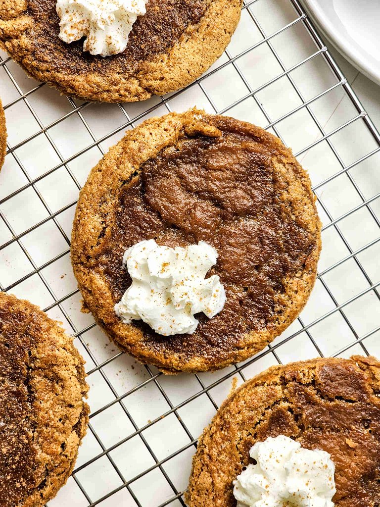 A close-up of pumpkin pie cookies topped with whipped cream and sprinkled spices, resting on a wire cooling rack.