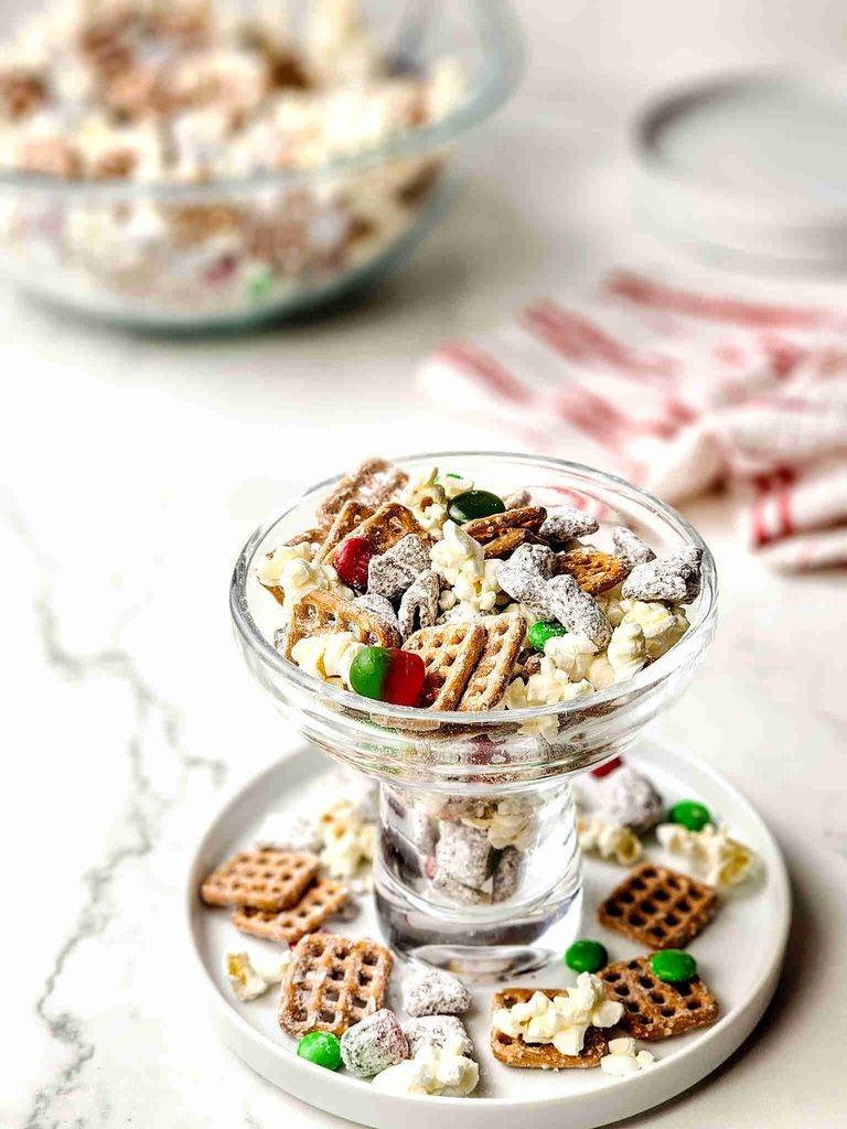 A glass footed bowl and small plate filled with reindeer munch, a snack mix of popcorn, pretzels, powdered sugar-coated cereal, and red and green candy-coated chocolates.