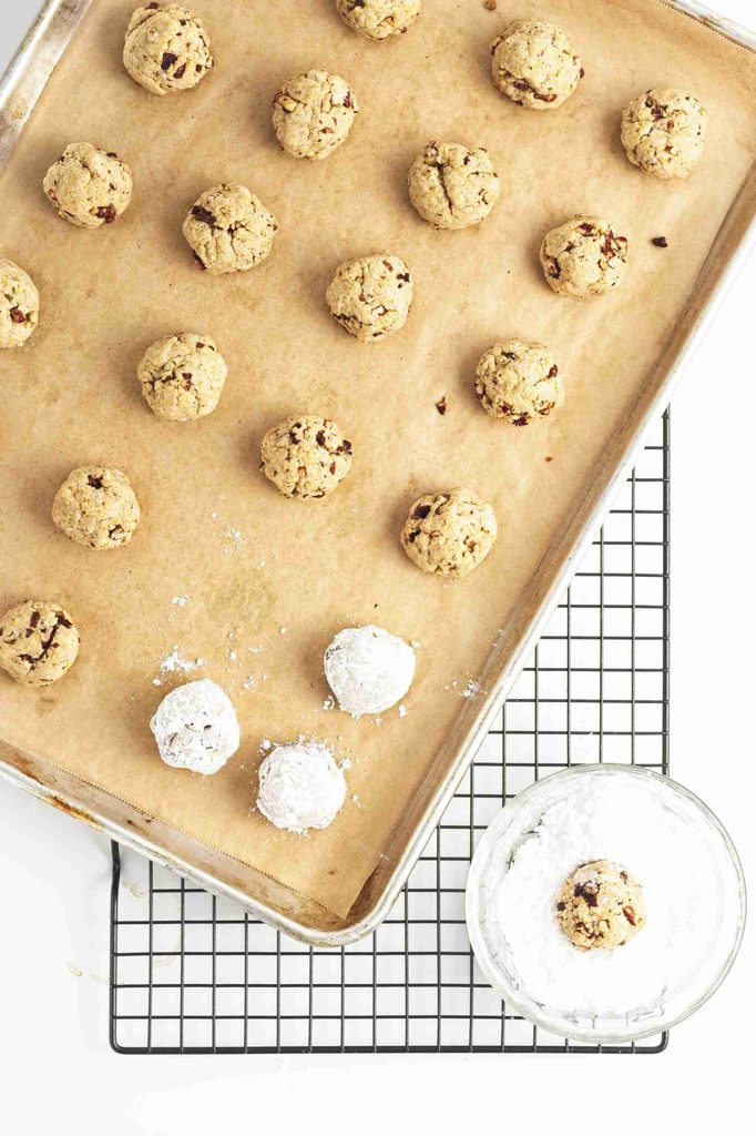 A baking sheet lined with parchment holds rows of unbaked pumpkin snowball cookies, some dusted in powdered sugar. Below, a wire rack and bowl of powdered sugar are visible.