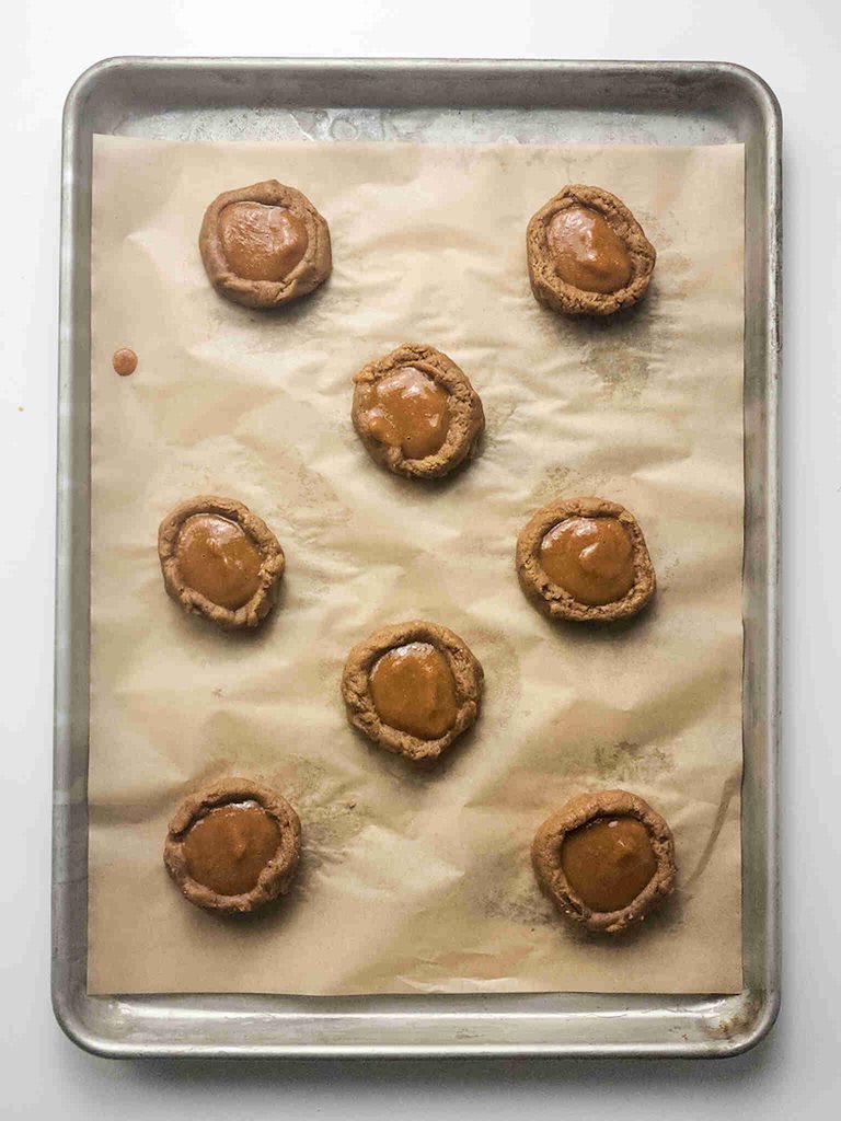 Nine round pumpkin pie filled cookies with caramel centers are spaced on a parchment-lined baking sheet. The pumpkin pie cookies are slightly cracked around the edges and arranged in an uneven pattern.