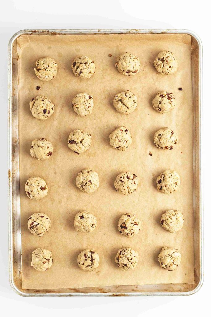A baking tray lined with parchment paper holds twenty balls of raw pumpkin snowball cookies, arranged in a grid pattern, ready to be baked.