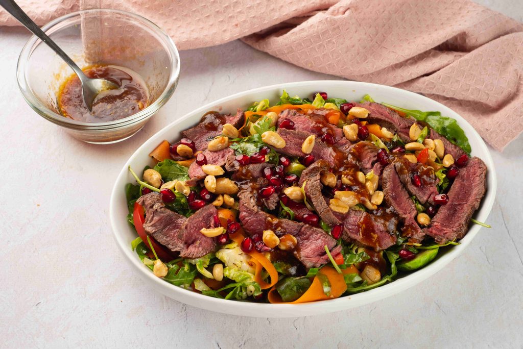 A fall steak salad of sliced beef, peanuts, pomegranate seeds, and greens in an oval dish, with a small bowl of dressing nearby.