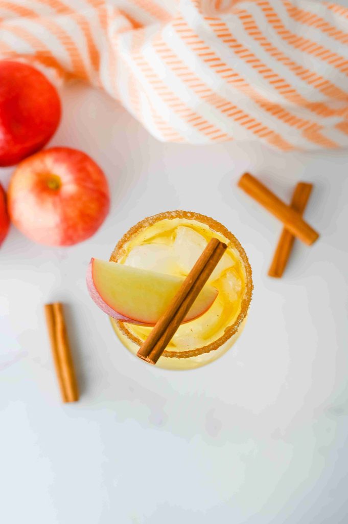 A top-down view of a Thanksgiving vodka cocktail in a sugar-rimmed glass, garnished with an apple slice and a cinnamon stick, surrounded by fresh apples and more cinnamon sticks.