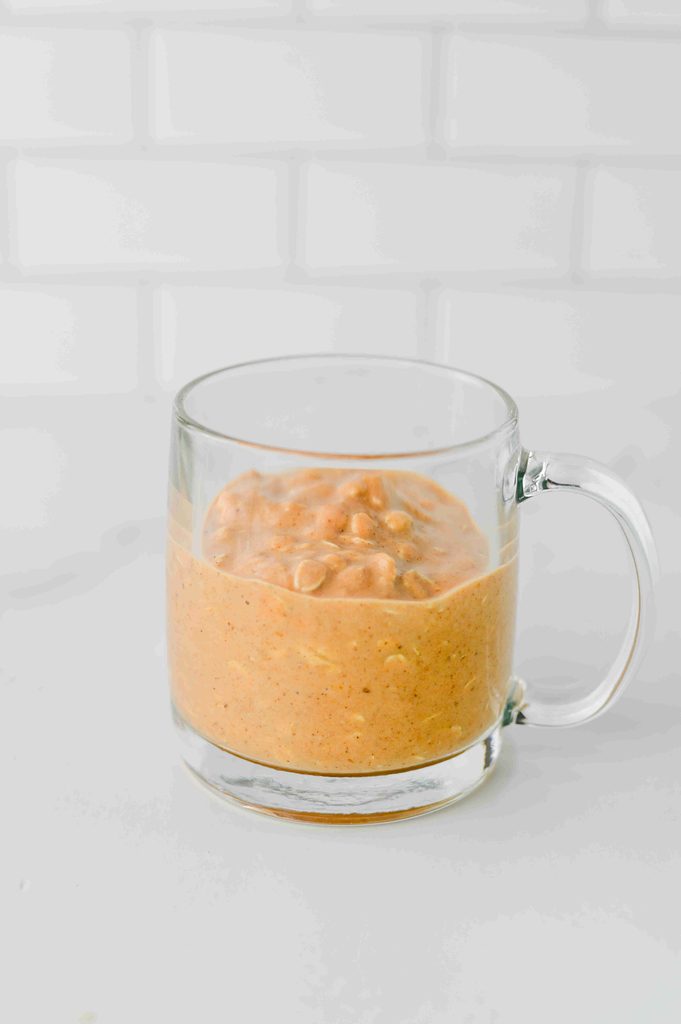 A glass mug filled with a thick, orange-colored blended high protein pumpkin overnight oats.