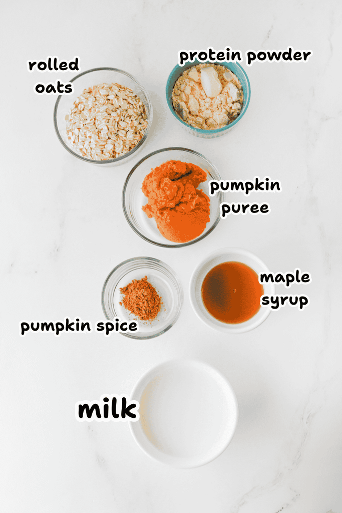Six small bowls arranged on a white surface, containing ingredients for high protein pumpkin overnight oats: rolled oats, protein powder, pumpkin puree, pumpkin spice, maple syrup, and milk.