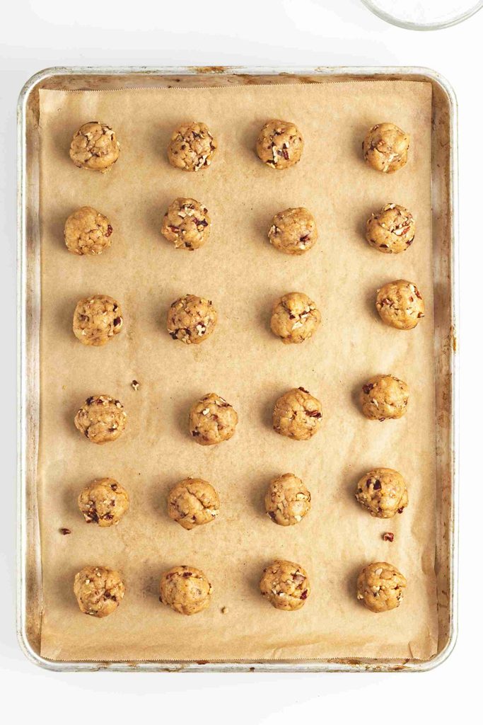 A baking sheet lined with parchment paper holds 24 evenly spaced balls of pumpkin snowball cookie dough, ready to be baked.