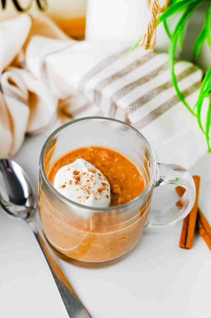 A glass mug of high protein pumpkin overnight oats topped with a dollop of whipped cream and cinnamon, beside a spoon and cinnamon sticks.