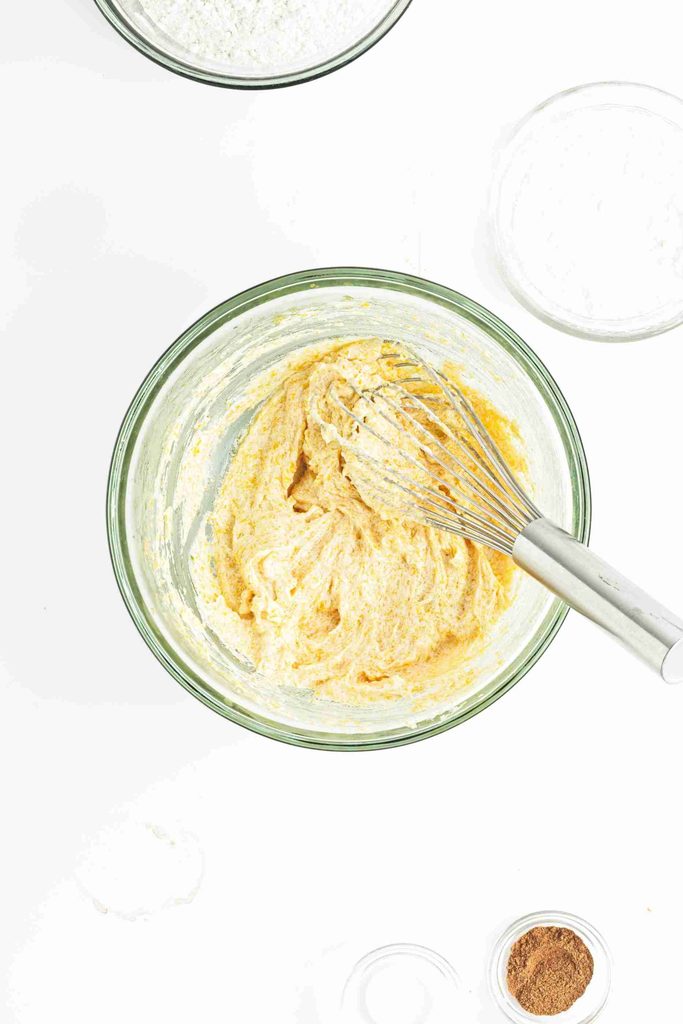A glass bowl filled with yellow batter for pumpkin snowball cookies is being mixed with a metal whisk. Surrounding the bowl are small, empty glass bowls and a small bowl with brown spice, all on a white surface.