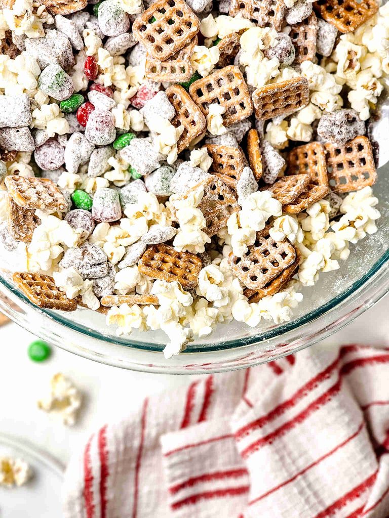 A clear glass bowl filled with reindeer munch, a festive snack mix of popcorn, pretzels, and powdered sugar-coated cereal with red and green candies.