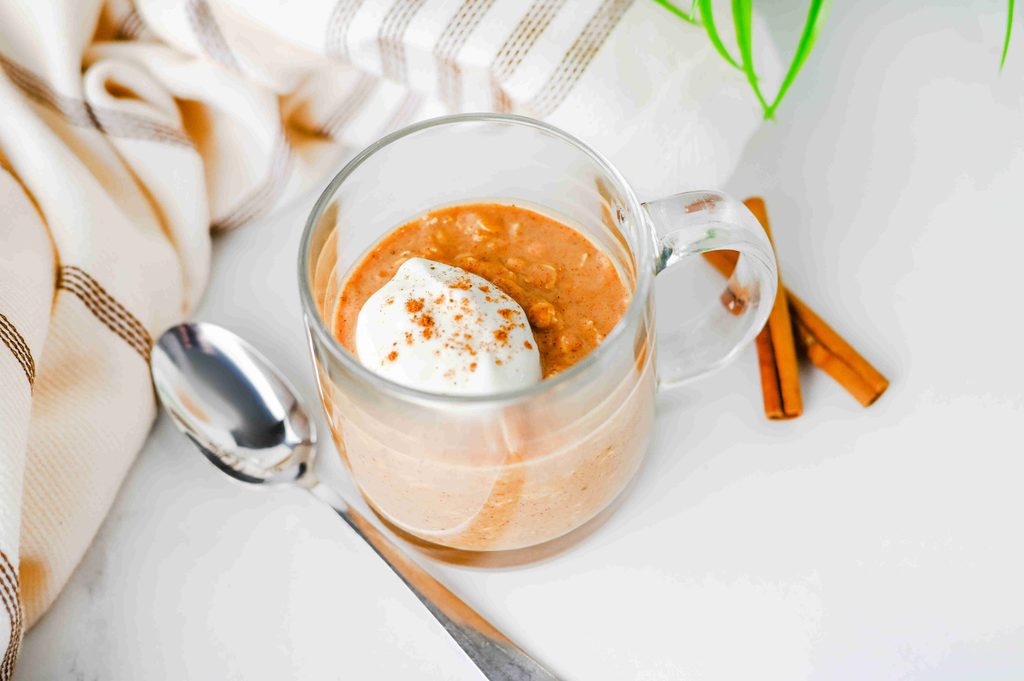 A glass mug of high protein pumpkin overnight oats topped with a dollop of cream and cinnamon, served with a spoon and cinnamon sticks.