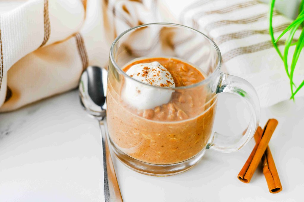 A glass mug filled with high protein pumpkin overnight oats, topped with whipped cream and a sprinkle of cinnamon, next to a spoon and cinnamon sticks.