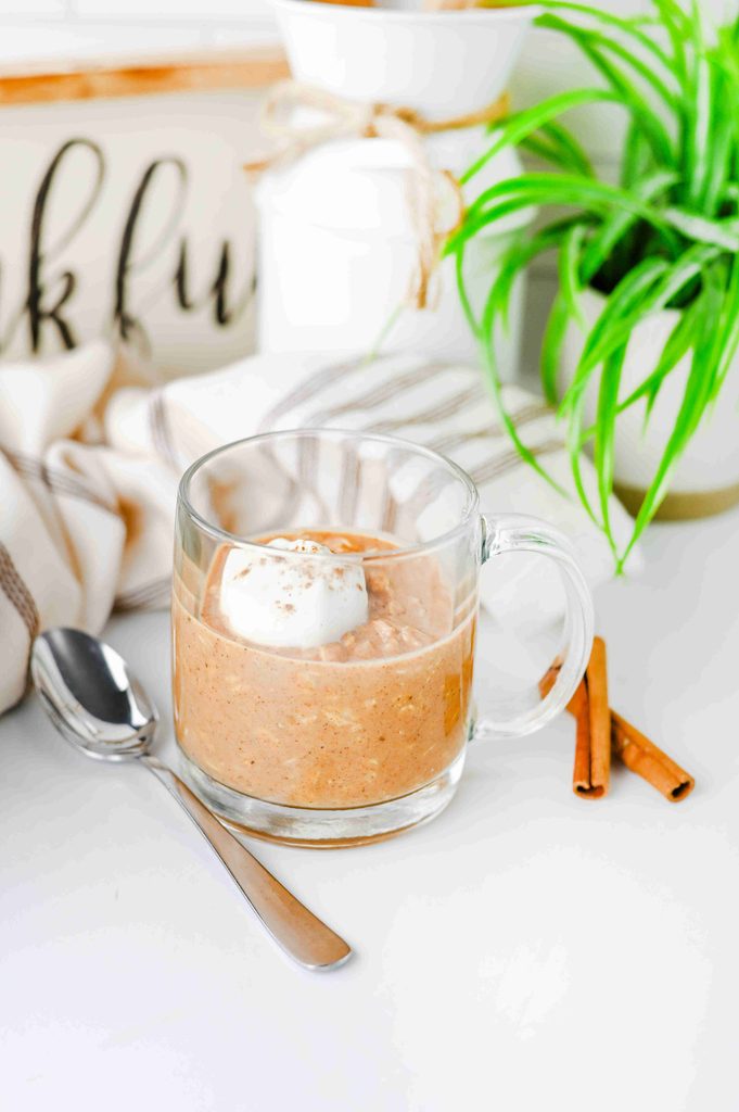 A glass mug filled with high protein pumpkin overnight oats topped with a dollop of whipped cream, next to a spoon and cinnamon sticks.