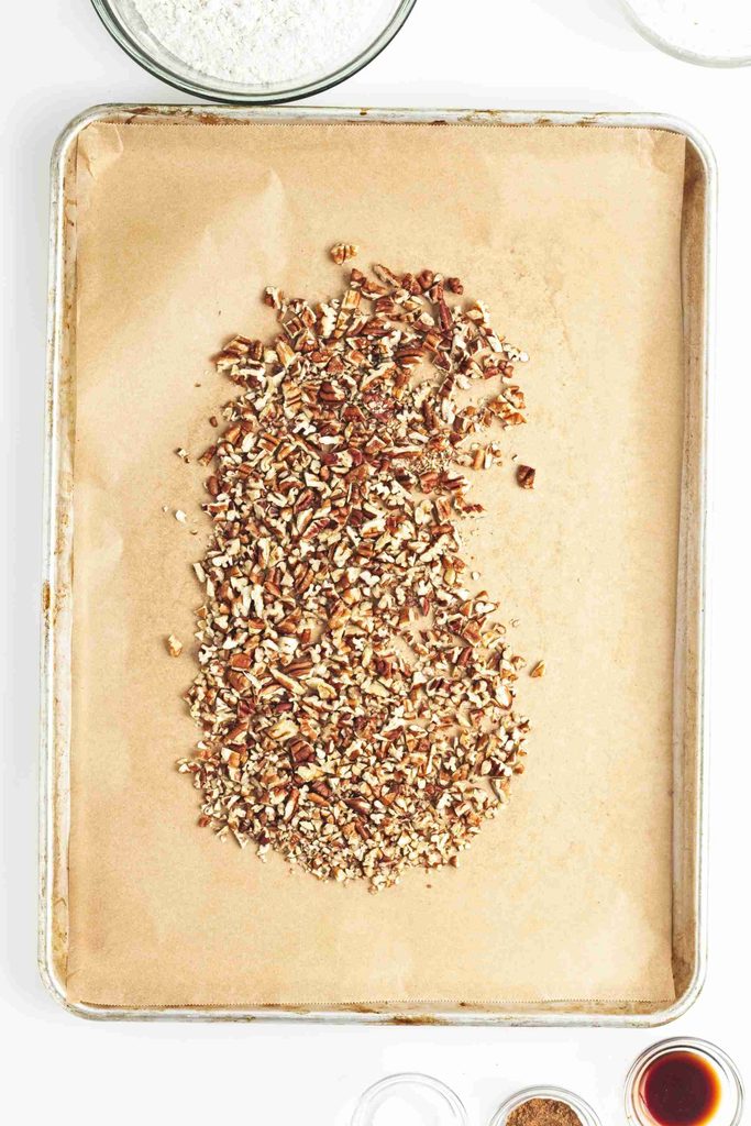 Chopped pecans spread on a parchment-lined baking sheet, ready for roasting—perfect for making vegan snowball cookies. Partial bowls of ingredients can be seen at the top and bottom edges of the image.