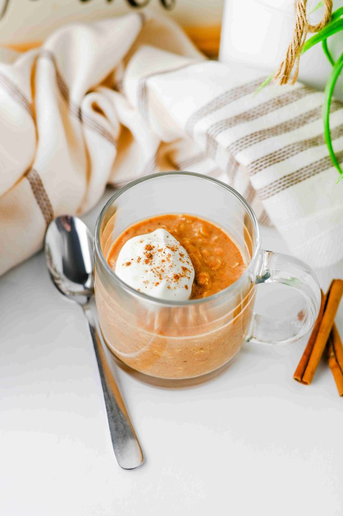 A glass mug filled with high protein pumpkin overnight oats topped with a dollop of whipped cream and cinnamon, served with a spoon and cinnamon sticks.