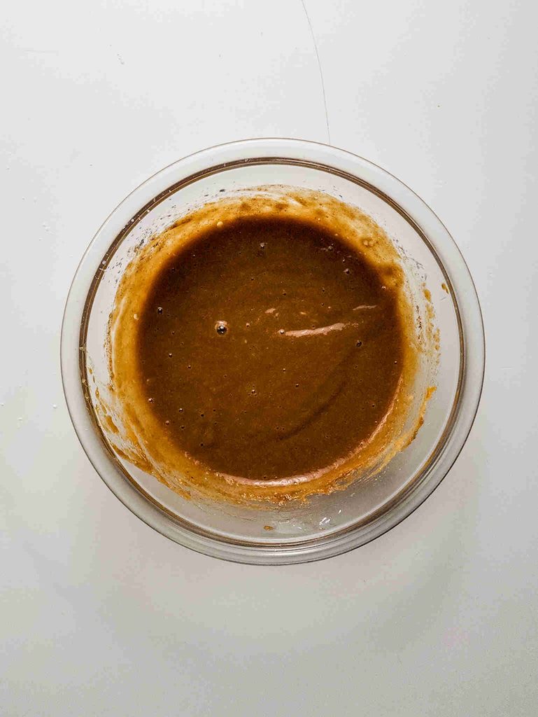 A clear glass bowl filled with thick, brown pumpkin pie cookie batter sits on a white surface. The smooth mixture, perfect for baking pumpkin pie cookies, has a few small bubbles on the surface.
