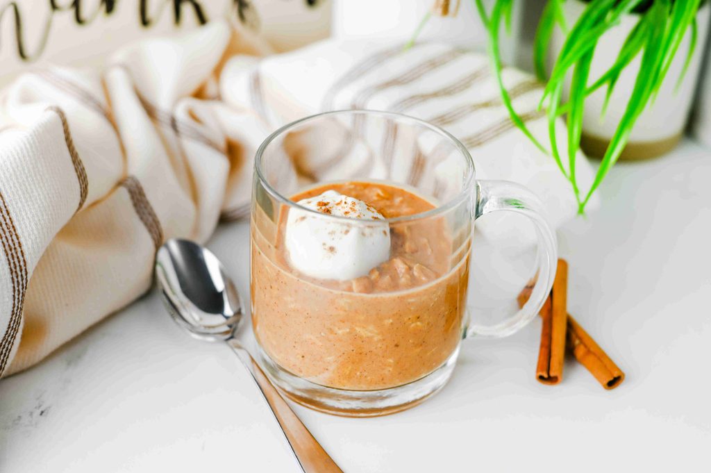 A clear glass mug filled with high protein pumpkin overnight oats topped with whipped cream and cinnamon, served with a spoon and cinnamon sticks.