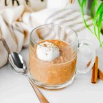 A clear glass mug filled with high protein pumpkin overnight oats topped with whipped cream and cinnamon, served with a spoon and cinnamon sticks.