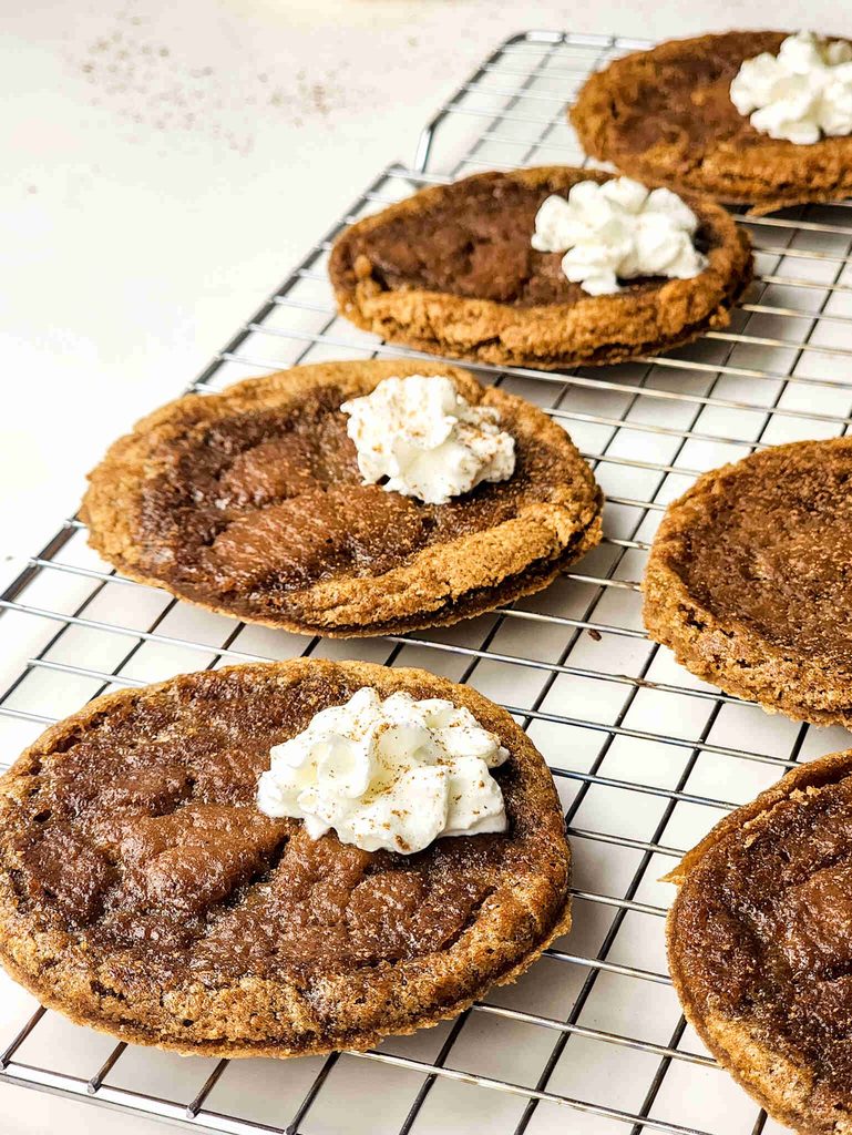 Six mini pumpkin pie tarts with custard filling and dollops of whipped cream are cooling on a wire rack. The tarts have a golden-brown crust, spiced filling, and a sprinkle of seasoning atop the whipped cream.